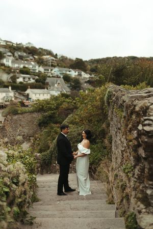 A couple stands on stone steps along a coastal hillside with white houses dotting the landscape in the background.
