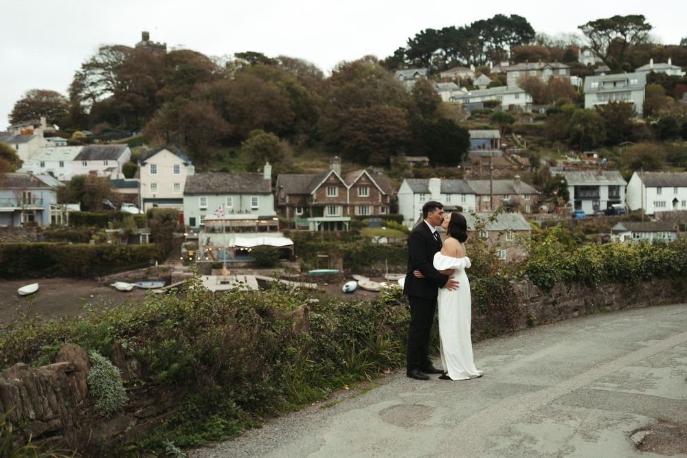 A couple poses in front of a coastal landscape with white houses and trees on a hillside overlooking the water.