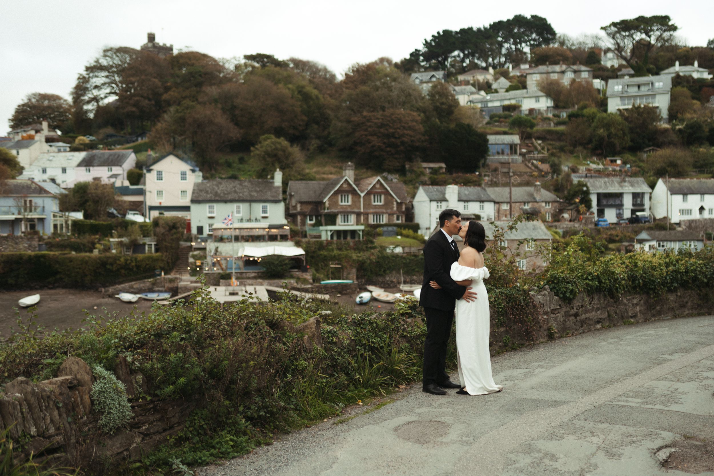 A couple poses in front of a coastal landscape with white houses and trees on a hillside overlooking the water.