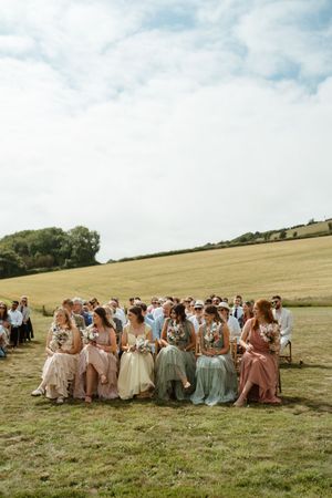 Wedding party in pastel dresses sits together on grassy hillside under cloudy sky for group photo.