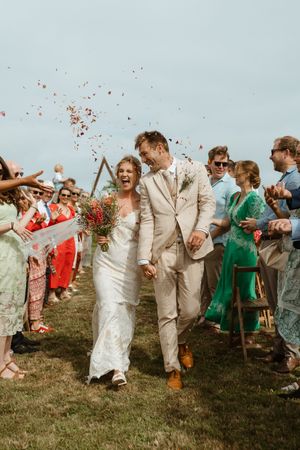 Wedding party guests throw confetti and celebrate as newlyweds walk down grassy hill in light-colored attire.