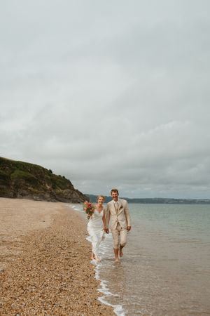 A couple walks along a scenic pebble beach shoreline on an overcast day with cliffs in the distance.
