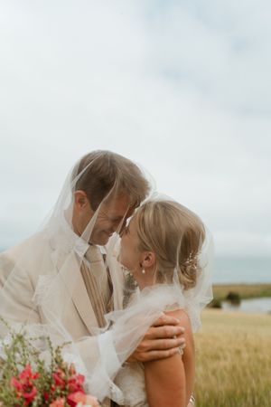 A romantic wedding couple embraces in a golden field with a veil flowing and red flowers near the ocean.
