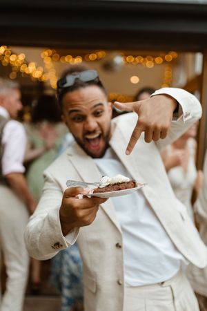 Someone in a white suit pointing excitedly at a slice of dessert at a celebration with bokeh lights in background.