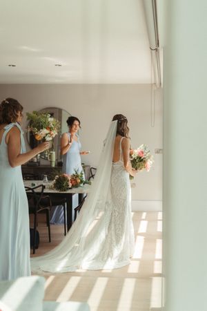 Bridesmaids in mint green dresses stand with bride wearing lace gown and veil in sunlit room with bouquets.