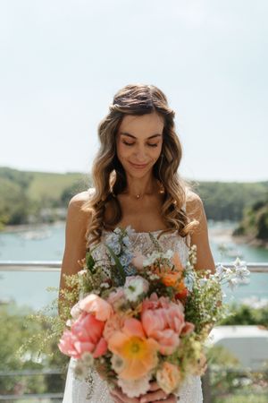 A bride in a white dress holds a vibrant bouquet of coral and peach flowers with a scenic water view in background.