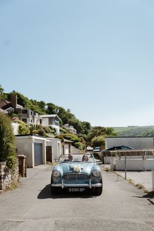 Classic vintage blue convertible car parked on a hillside road with coastal houses and clear blue sky in the background.