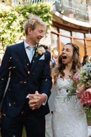 Joyful newlyweds share candid laughter and emotions during outdoor garden wedding celebration with fluttering confetti.
