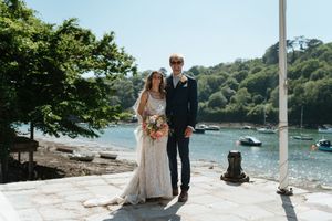 Newlywed couple stands together on stone pier overlooking scenic coastal bay with boats and green hills in background.