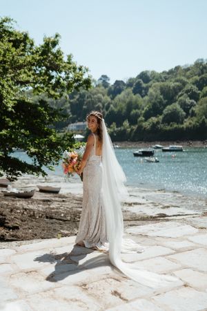 A bride in a fitted lace gown and long veil holds a bouquet by a scenic waterfront with moored boats and trees.