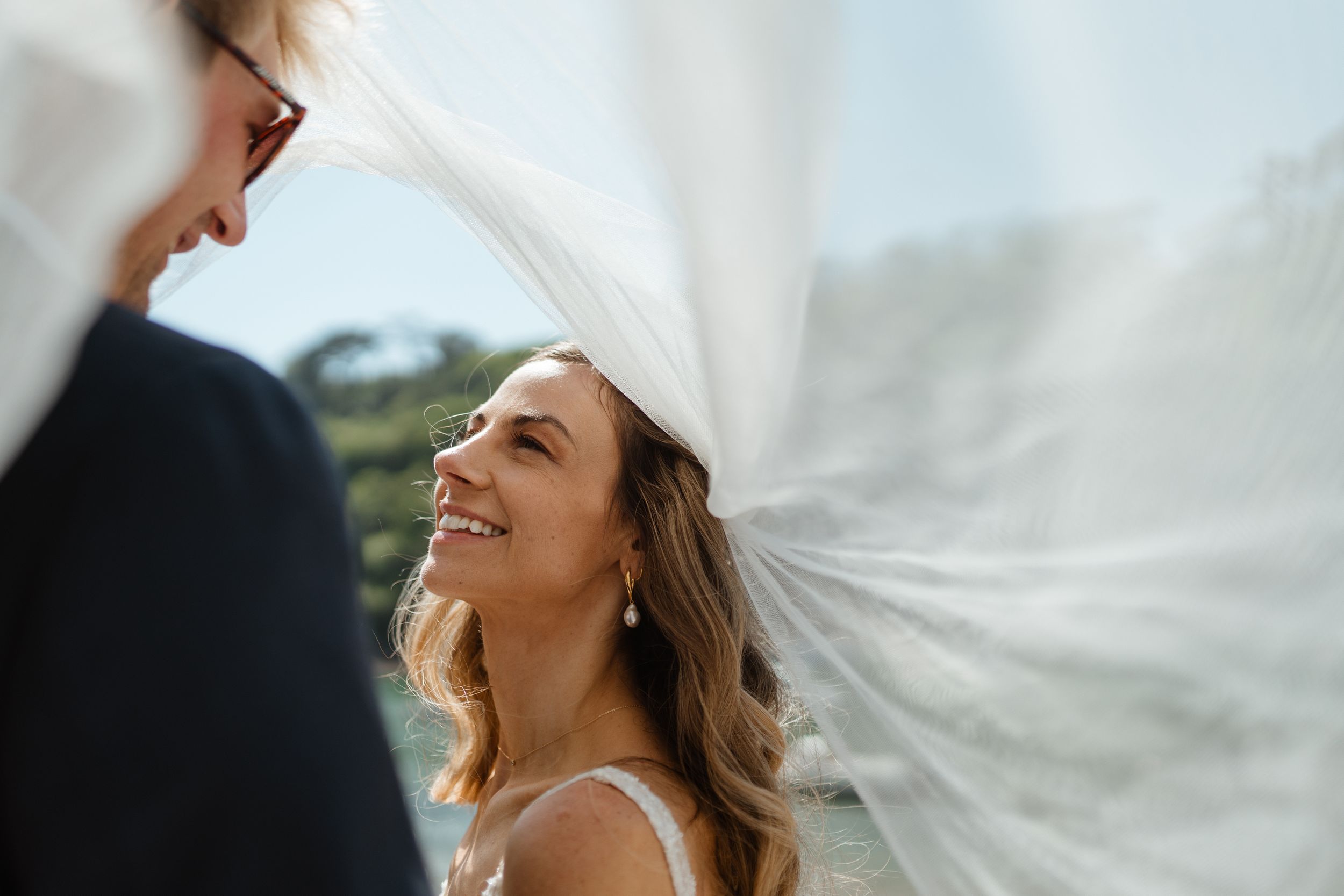 A bride smiles joyfully under a flowing veil during an outdoor wedding ceremony.