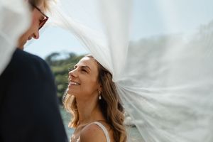 A bride smiles joyfully under a flowing veil during an outdoor wedding ceremony.