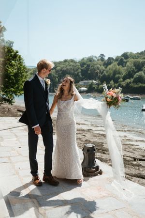 A bride and groom share a joyful moment together on a waterfront stone patio with a flowing veil and floral bouquet.