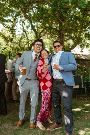 Wedding guests share candid moments and laughter in a casual outdoor setting with trees and dappled sunlight.