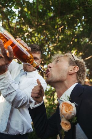A groomsman in a suit pours alcohol into the groom's mouth from a bottle at an outdoor wedding celebration.