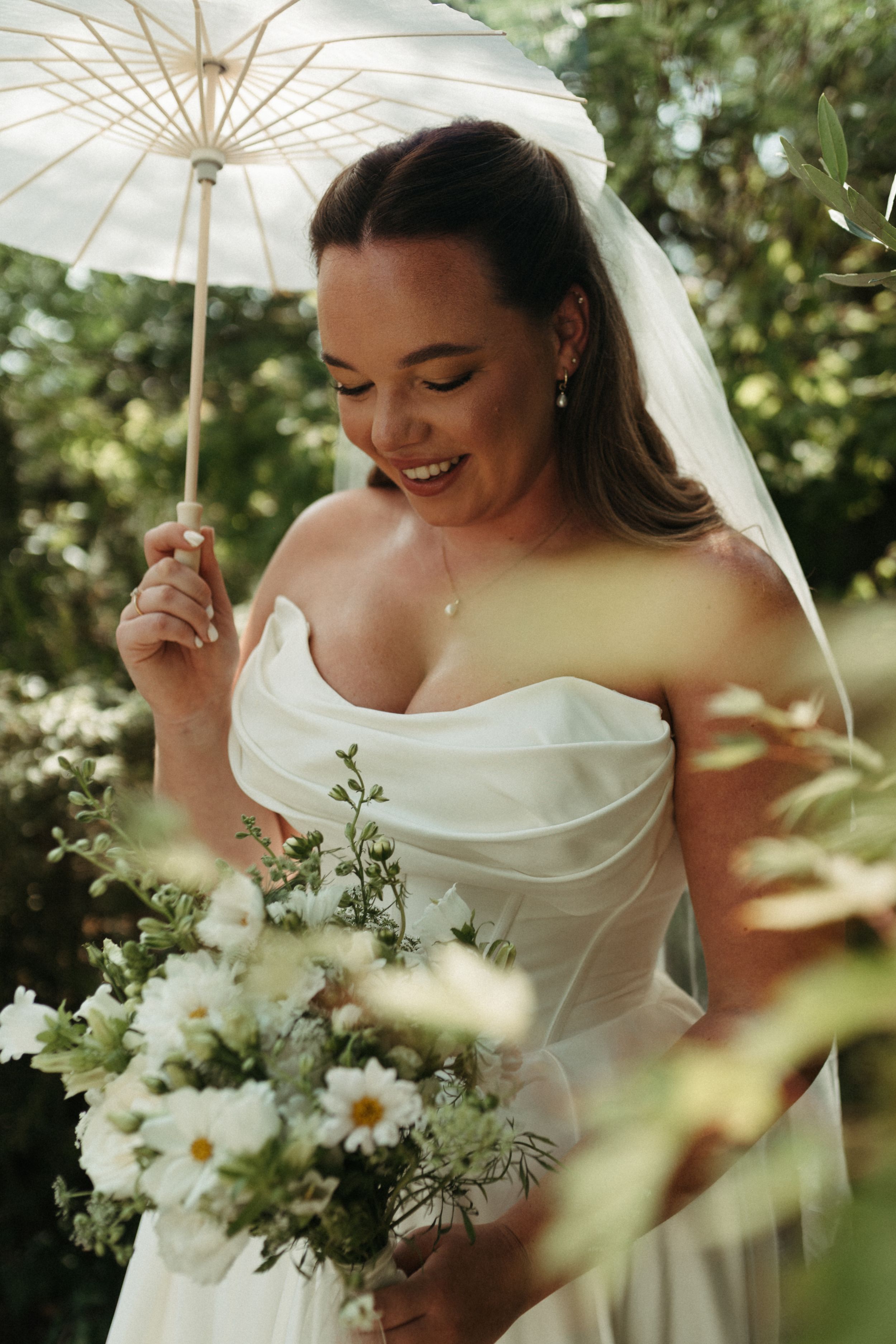 Smiling bride in strapless white gown holds daisy bouquet and parasol umbrella outdoors in lush garden setting.