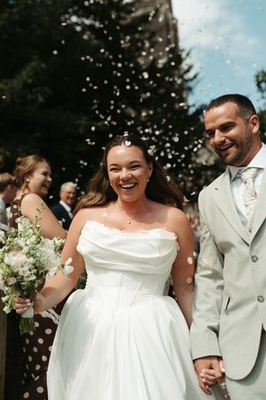 A joyful bride in a strapless white gown and groom in a light suit walk through confetti after their wedding ceremony.