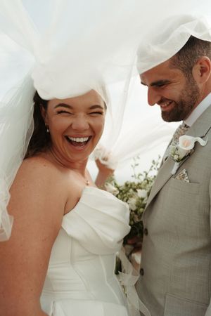 Bride laughing joyfully with groom at wedding, veil blowing in wind, white dress and grey suit.