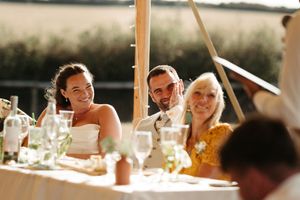 Happy bride and groom smiling at wedding reception table with guests under outdoor marquee with fairy lights.