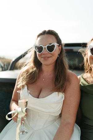 Bride in white strapless dress and heart-shaped sunglasses holding champagne glass outdoors on sunny day.