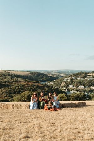 Group of women and children sitting on hay bales in a golden field with scenic valley and village views behind them.