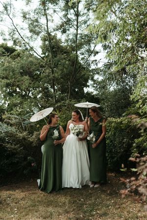 Bride in white gown with two bridesmaids in green dresses holding white parasols, posing in a lush garden setting.
