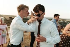 Two men in white dress shirts share a drink outdoors at a wedding reception on a sunny day.