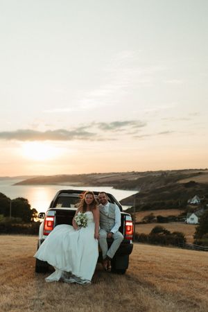 Bride and groom sitting on truck tailgate at sunset overlooking coastal landscape on wedding day.