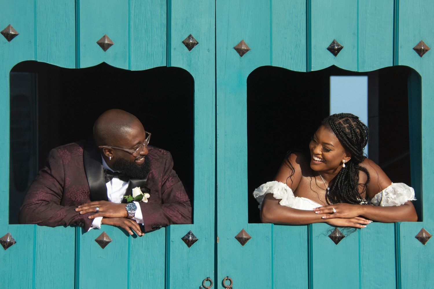 Bride and groom lean through turquoise wooden door windows sharing romantic moment at wedding venue.
