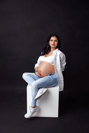 Maternity photo of woman in white top and jeans sitting on white cube against dark background.