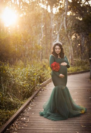 A maternity photo featuring a green tulle gown and red bouquet on a wooden boardwalk at sunset.