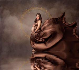 Artistic maternity portrait in flowing brown fabric with golden halo effect against dark backdrop.