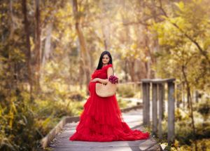Maternity portrait on wooden bridge with dramatic red tulle gown in autumn setting.