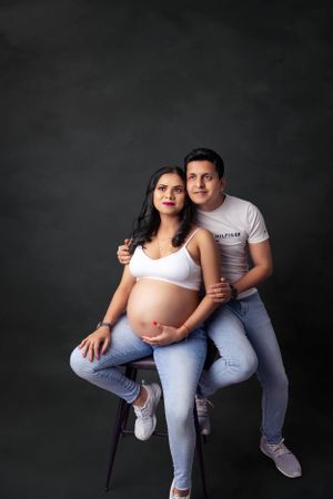 Studio maternity photo with casual denim and white attire against dark background.
