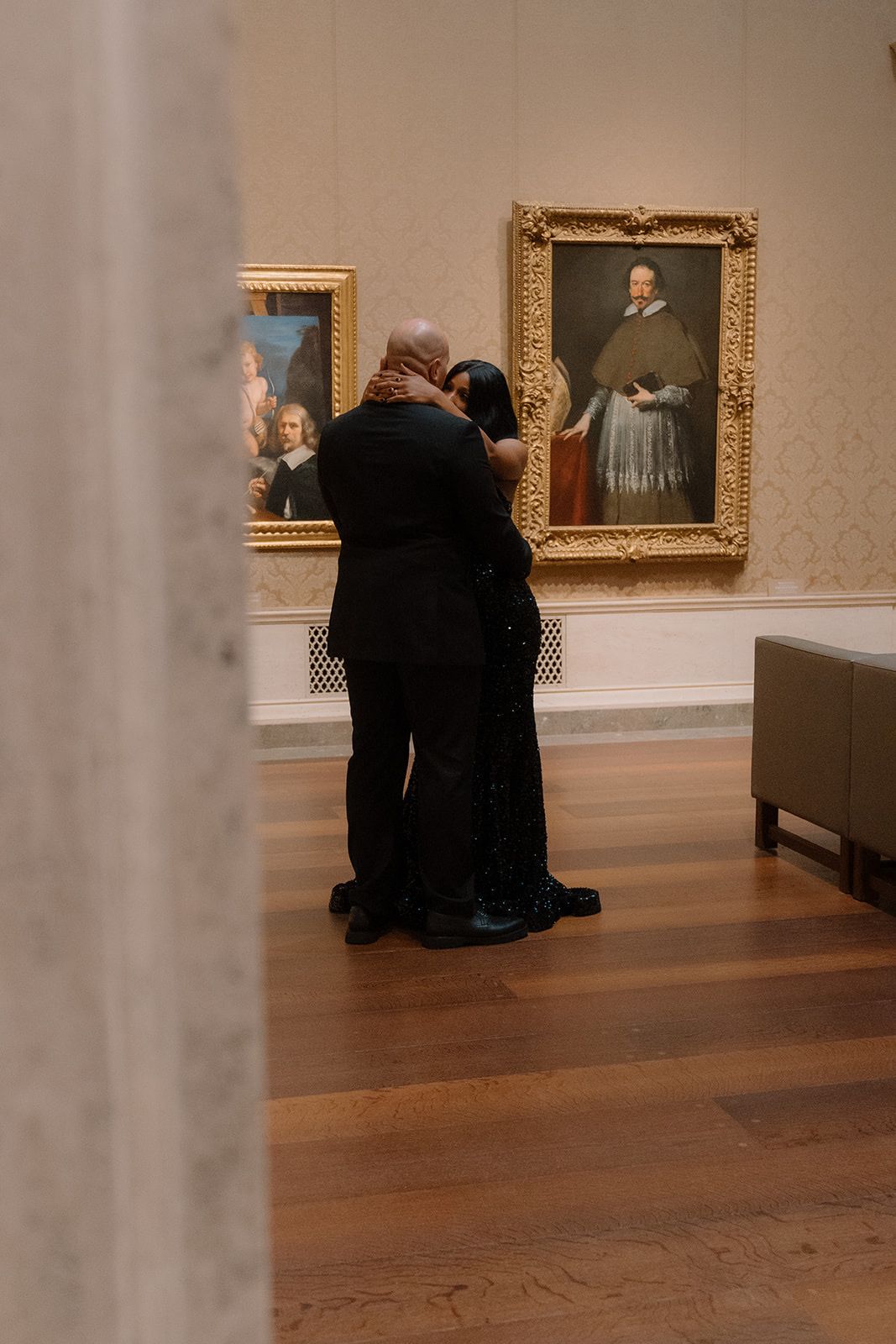 Two people embrace while looking at classical portrait paintings in an art museum gallery with wooden floors during an engagement session