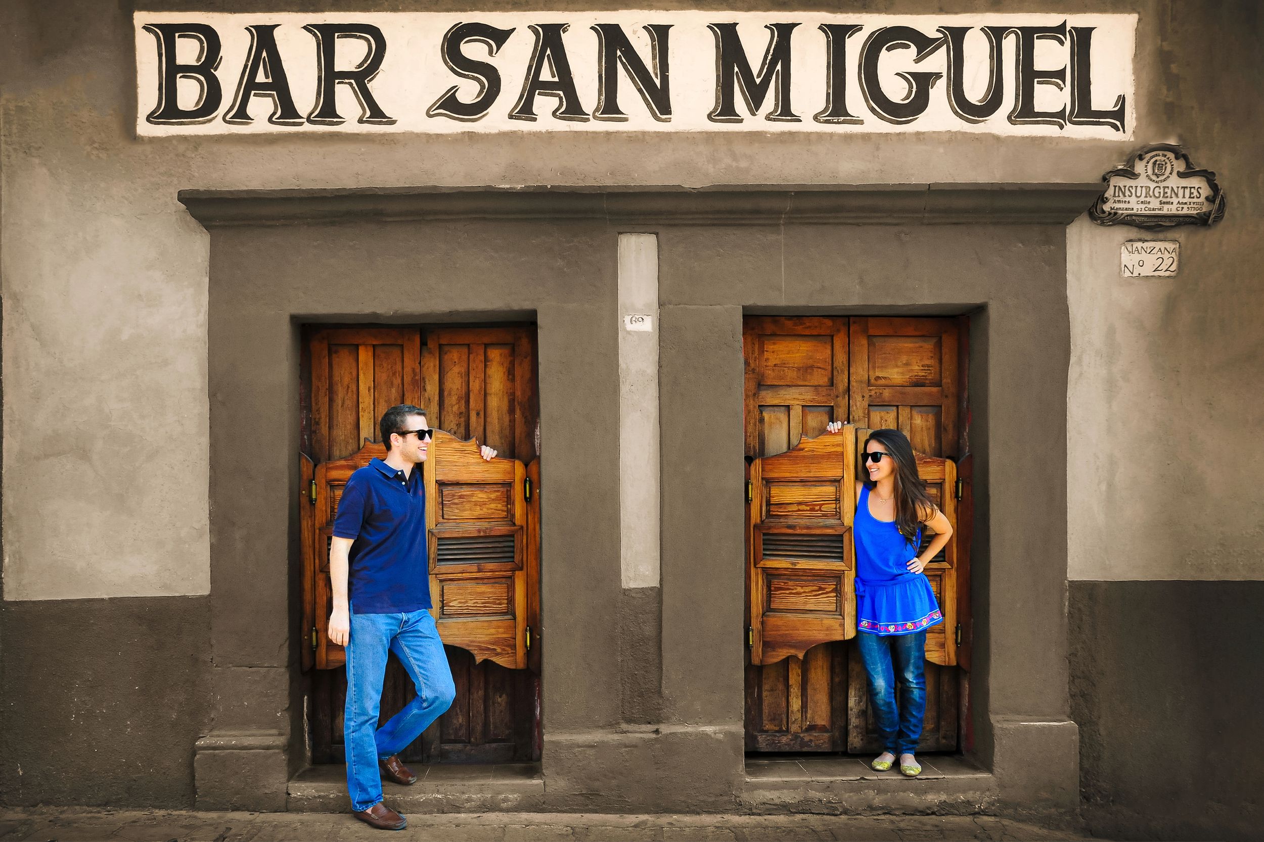 Two people in blue outfits stand in wooden doorways of Bar San Miguel.