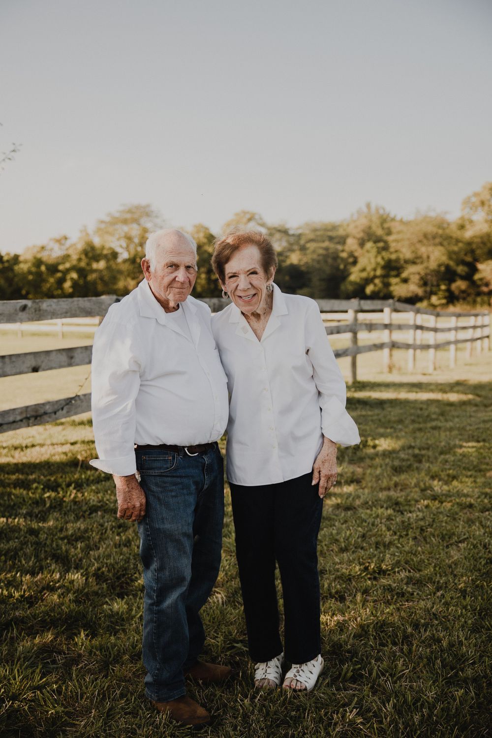 Senior couple in white shirts standing together on grassy field with fence in background.