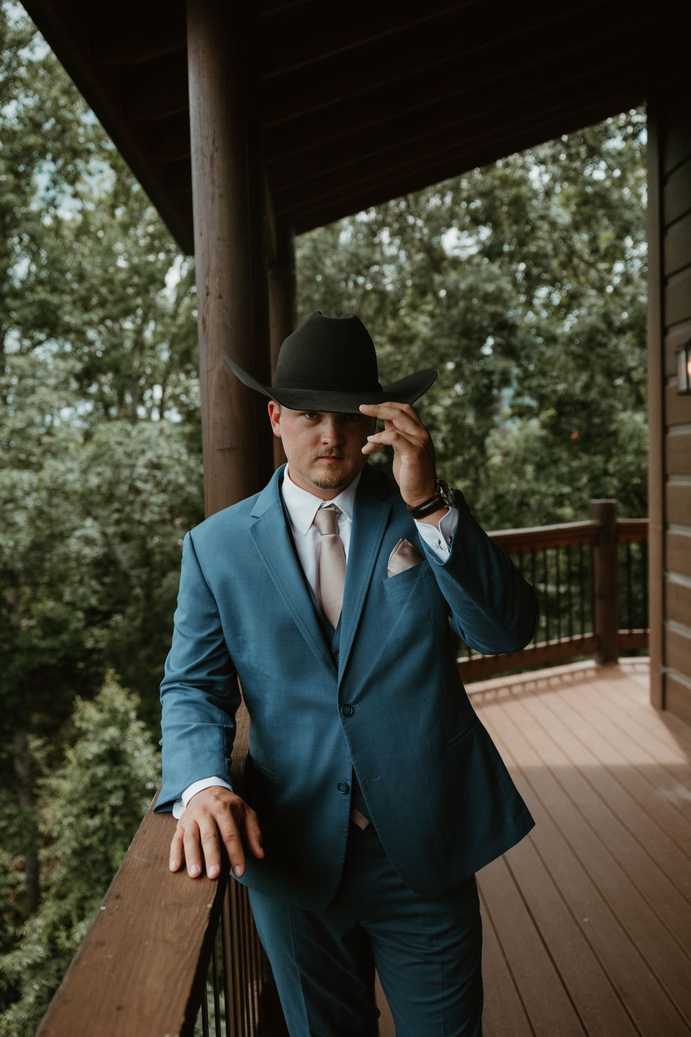 Groom in blue suit and baseball cap poses on wooden deck railing.