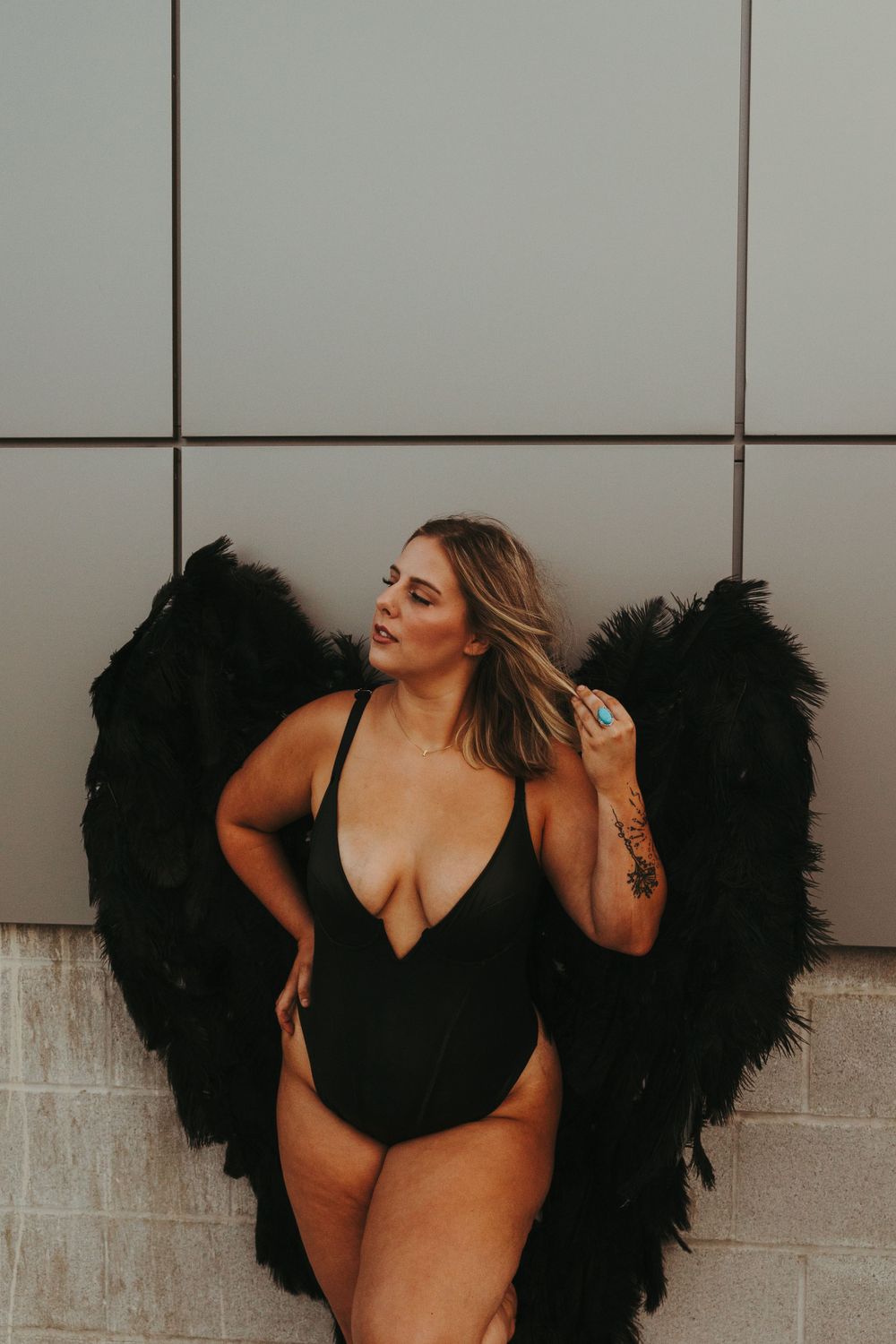 Woman in black swimsuit poses against white wall with dramatic dark angel wings.
