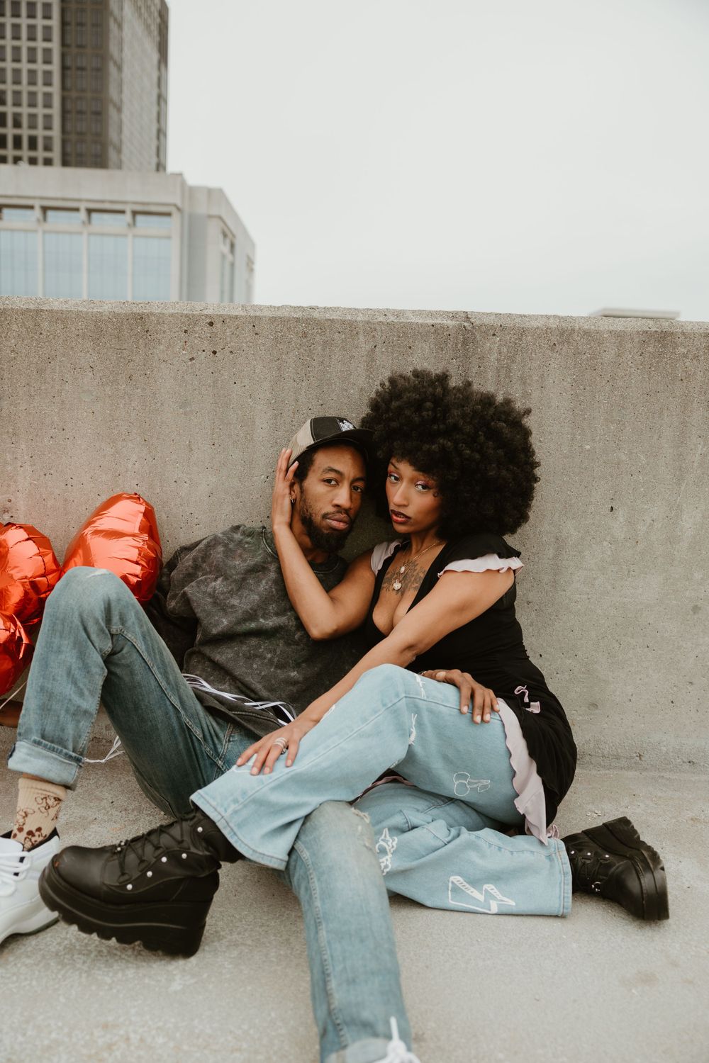 Couple lounging against concrete wall with red heart balloons in urban setting.