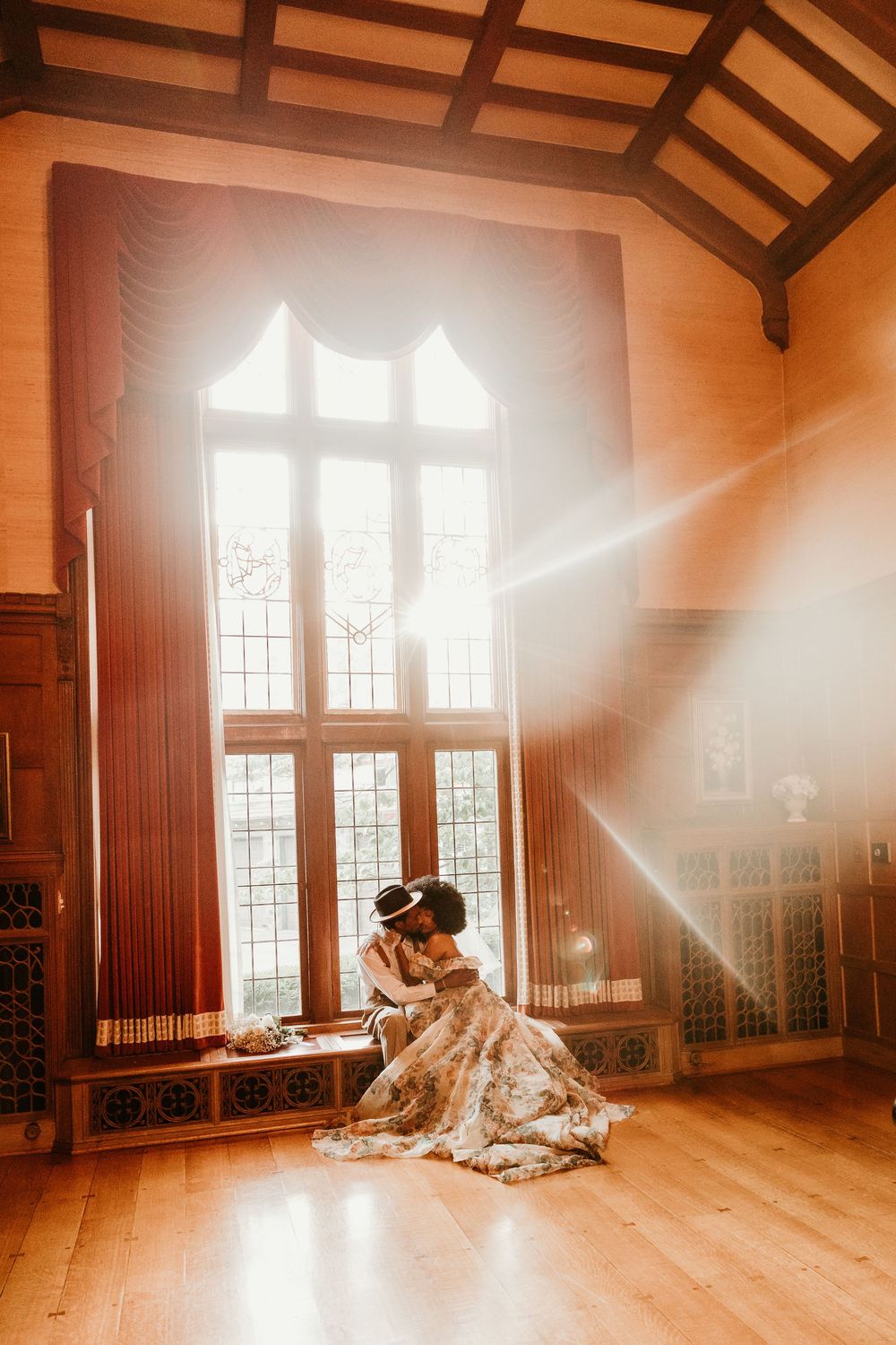 Sunlight streams through gothic window onto couple embracing in vintage dress on wooden floor of historic building.