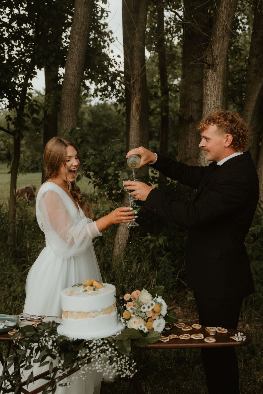 Couple shares playful cake-smashing moment during rustic outdoor wedding reception in wooded setting.