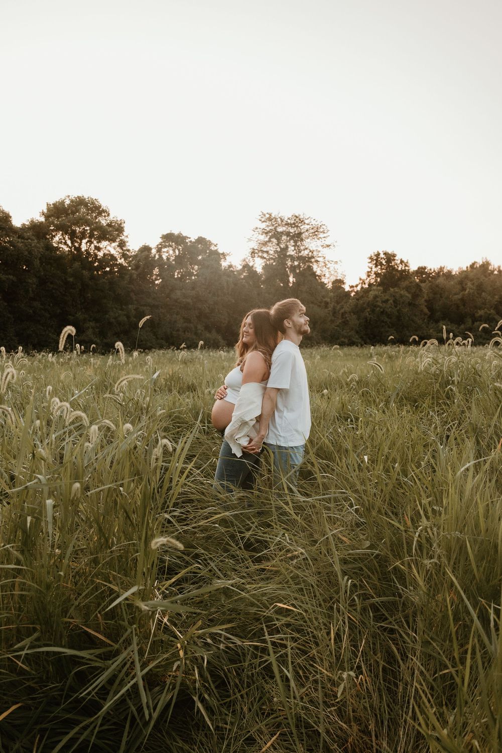 A couple stands close together in a tall grass field at sunset with trees silhouetted in the background.