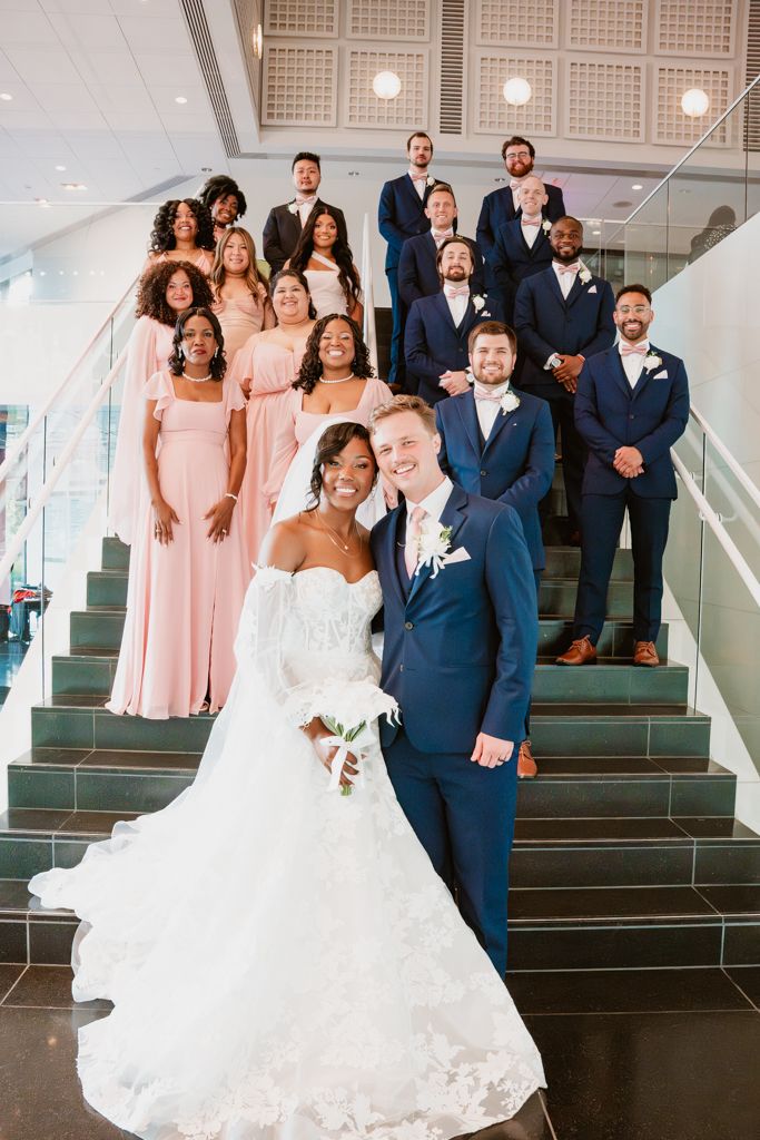 Wedding party poses on modern staircase with bridesmaids in pink dresses and groomsmen in navy suits.