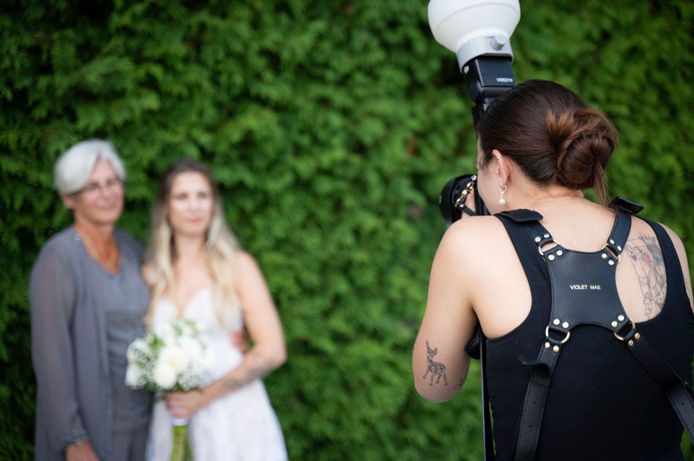 A photographer with a light modifier capturing family photos for a wedding against a lush green hedge background.