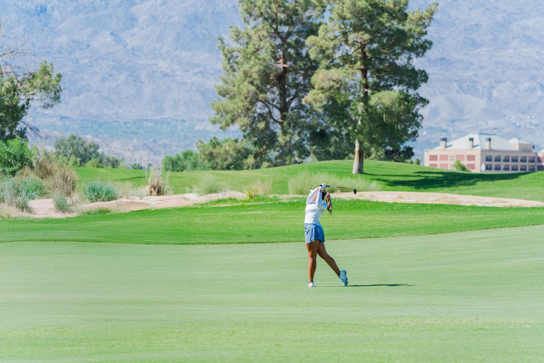 A golfer walks across a lush green golf course with pine trees and mountains in the background.