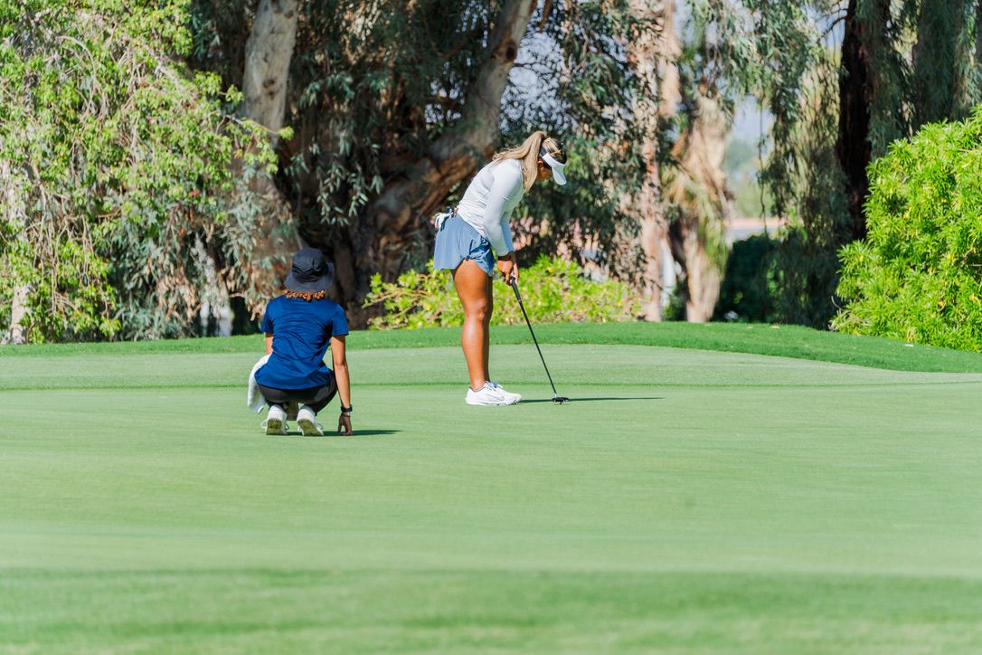 Two golfers on a green putting course with one lining up a shot while the other analyzes the break.