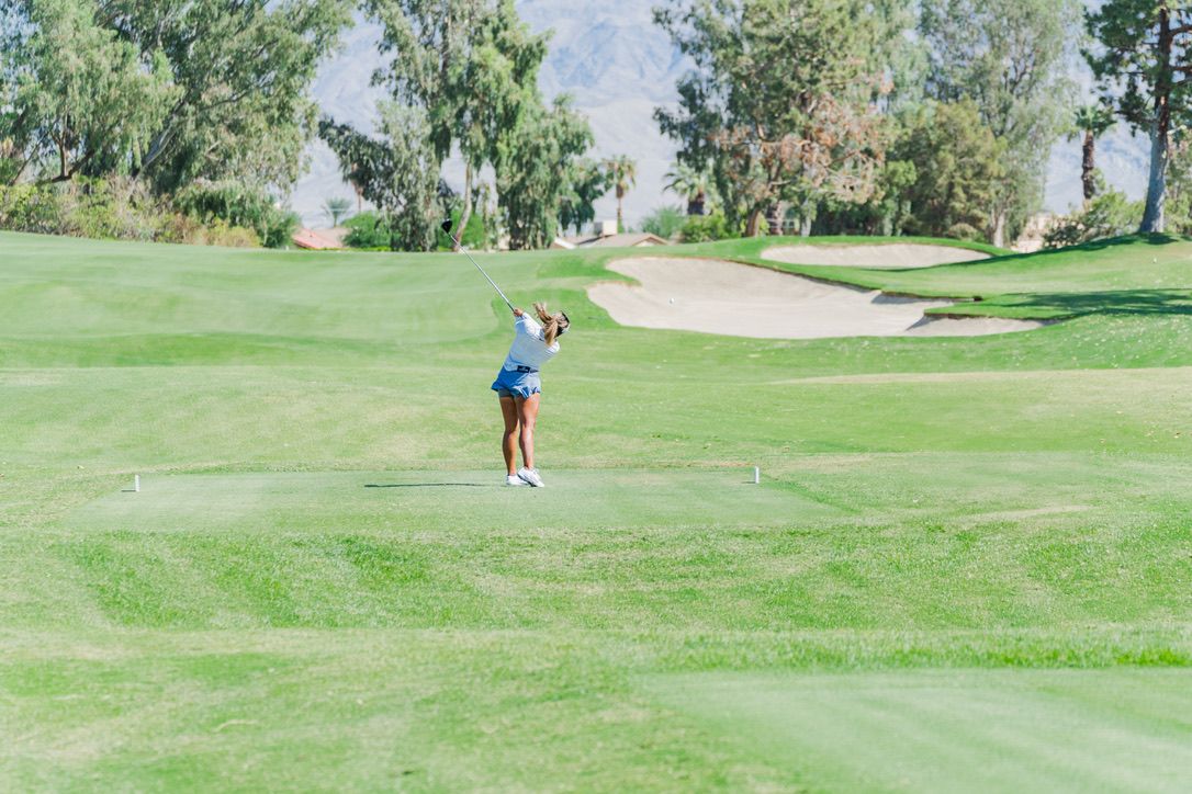 A golfer takes a shot on a well-manicured golf course with sand bunkers and willow trees in the background.