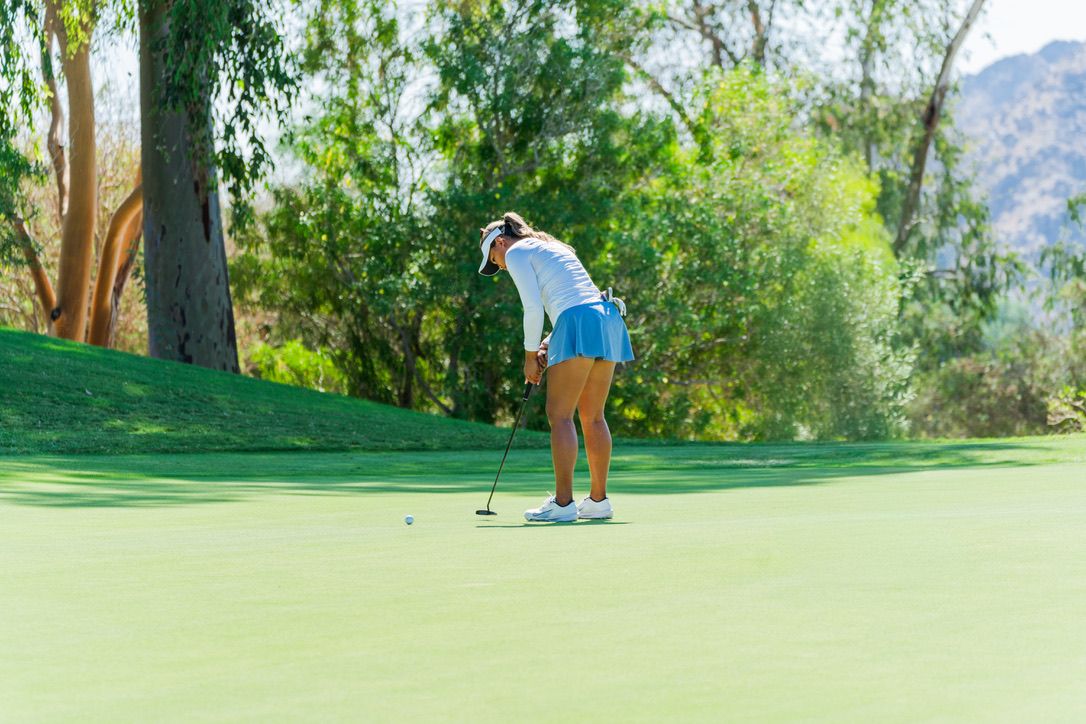 Golfer in blue attire putting on a pristine green golf course surrounded by trees and mountain scenery.