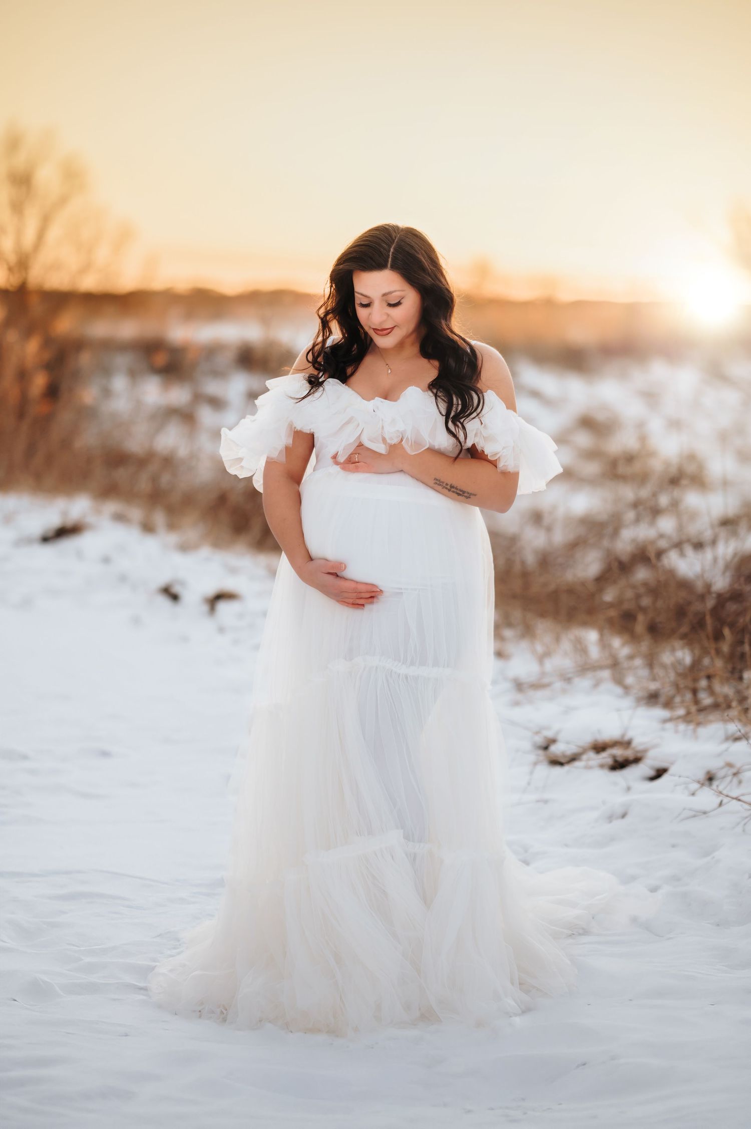 Dreamy winter maternity photo featuring flowing white dress against snow covered landscape at sunset.